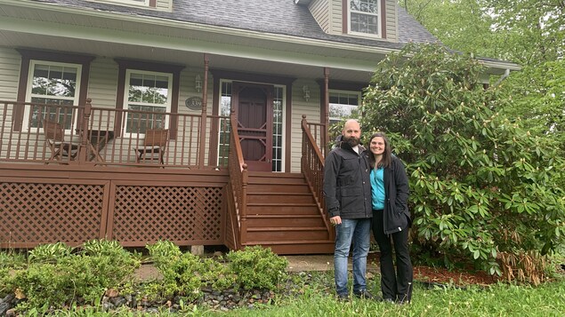 Un homme et une femme sourient à la caméra, devant leur maison,  l'extérieur.