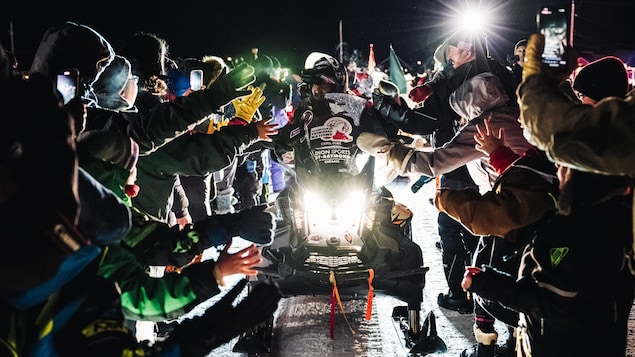 Un homme en motoneige, de nuit, avec beaucoup de monde autour qui tend la main.