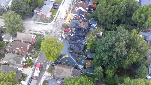 Photo des décombres d'une maison complètement démolie et de maisons adjacentes aux toits brûlés ou endommagés; un jet d'eau arrose l'un de ces toits.