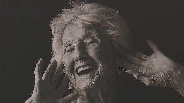 Une photo en noir et blanc d'une femme âgée en train de rire. Elle a ses deux mains près de son visage.