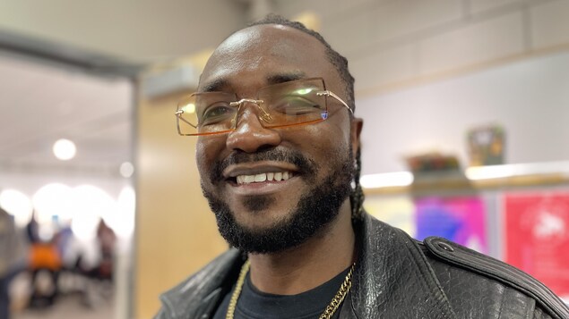 Ezoman porte une grosse chaîne au cou et des lunettes. Il sourit. 