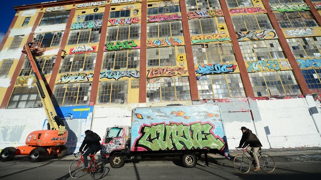Des cyclistes passent devant l'édifice 5Pointz, en partie recouvert de peinture blanche. Des cyclistes passent devant l'édifice 5Pointz, en partie recouvert de peinture blanche.