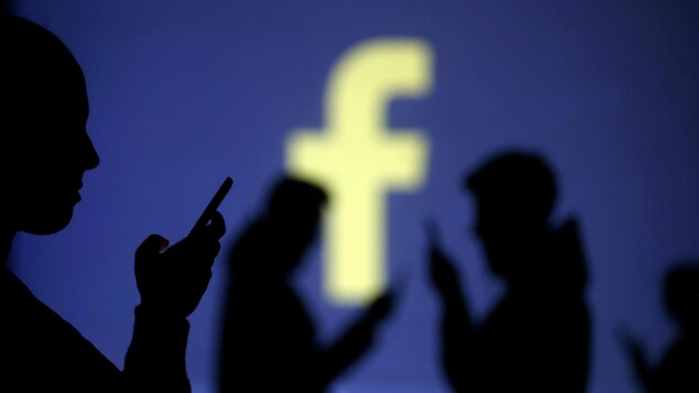 Des silhouettes de personnes qui utilisent des téléphones intelligents sont visibles devant un grand écran qui affiche le logo de Facebook.
