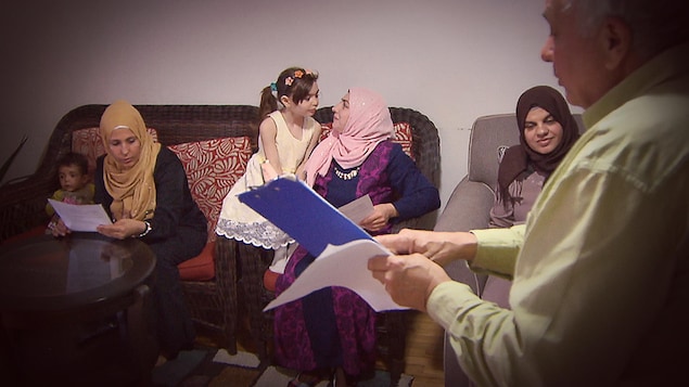 Un homme tiens des documents en main, trois femmes sont assises sur un canapé et elles l'écoutent parler