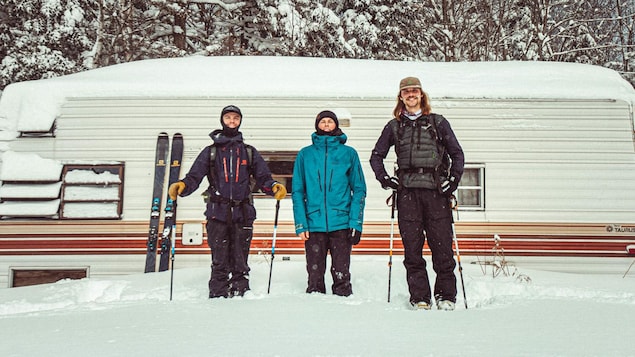 Trois jeunes hommes en tenue de ski sourient devant leur caravane