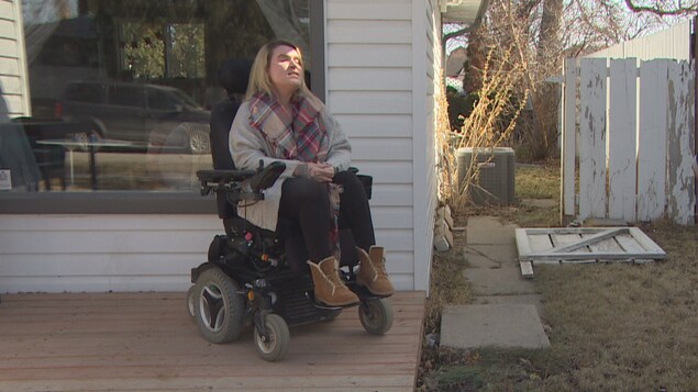 Une femme en fauteuil roulant est devant sa maison et parle à une journaliste.