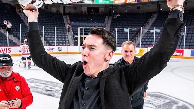 Sur la glace d'un aréna, un jeune homme soulève, victorieux, un grand trophée.