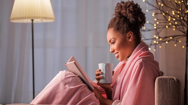Une jeune femme lit un livre, assise dans un sofa avec une tasse dans la main.