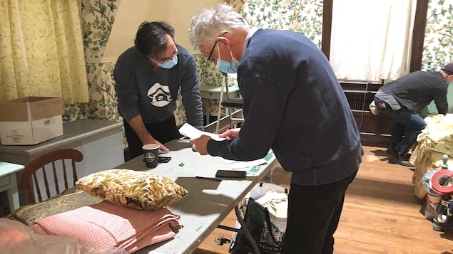 Deux hommes, face à face, portant un masque médical, travaillent au-dessus d’une table.