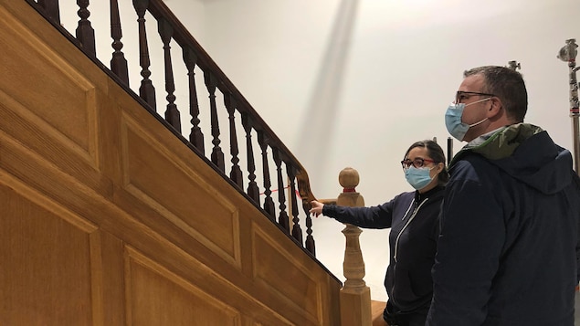 À droite, un homme et une femme qui portent un masque médical sont au pied d’un escalier en bois.