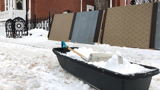 Dans la rue, à l’avant-plan, un traîneau en plastique est rempli de neige. Une pelle a été laissée sur le dessus. À l’arrière-plan, adossé à la clôture de fonte délimitant la cour de la maison, les panneaux reproduisant l’entrée intérieure de la maison.