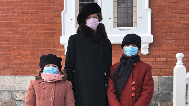 Hélène Florent, debout entre deux fillettes en âge de fréquenter une école primaire, est devant la maison de la rue des Ursulines. Les fillettes ont chacune un vieux sac de cuir à la main. Les trois portent un masque médical.