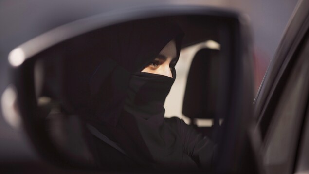 Dans un rétroviseur, le visage d'une femme au volant d'une voiture.