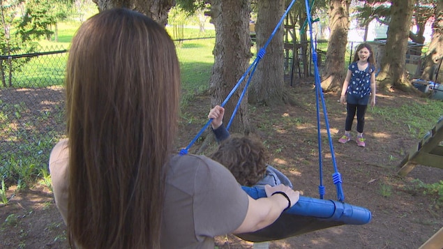 Une femme pousse un enfant sur une balançoire. Une autre enfant est debout de l'autre côté de la balançoire.
