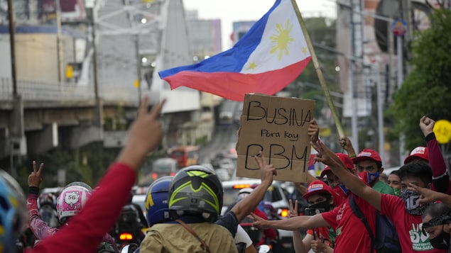 Mga tao na may hawak na bandila ng Pilpinas at isang karatula na nagsasabing 'Busina para kay BBM.'