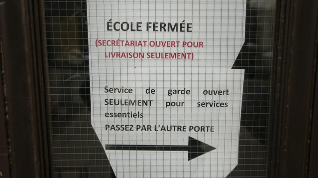 On peut lire sur une feuille de papier collée dans une fenêtre de porte : École fermée (secrétariat ouvert pour livraison seulement), Service de garde ouvert SEULEMENT pour services essentiels, Passez par l'autre porte.