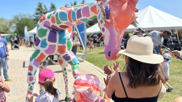 Trois fillettes tendent les mains vers une marionnette grandeur nature d'une giraffe à l'extérieur un jour d'été.