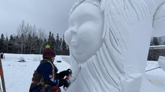 Une personne sculpte une visage en neige géant avec une scie mécanique.