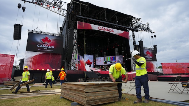 Des travailleurs transportent des planches de bois devant une scène aux couleurs du drapeau canadien.
