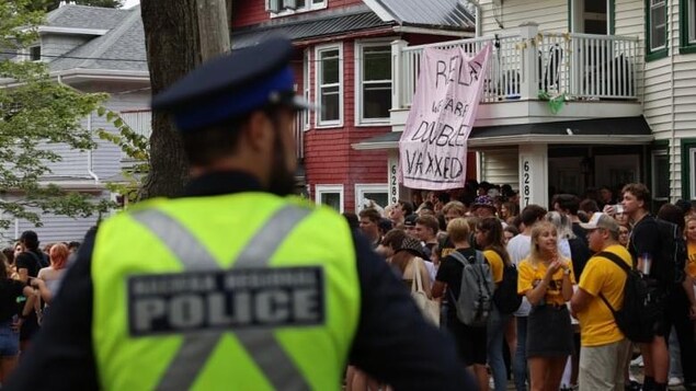 Une police regarde une foule de jeunes près d'une maison dans un quartier résidentiel, où une pancarte affiche « relaxez, nous sommes doublement vaccinés ».