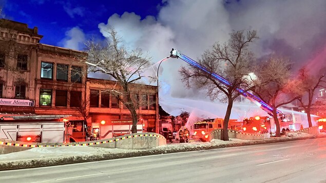 Quatre camions de pompiers arrosent un bâtiment où de la fumée sort.