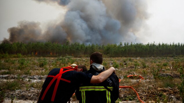 Des pompiers s'enlacent, exténués, devant un feu de forêt.