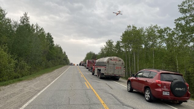 Un avion-citerne passe au-dessus d'une route sur laquelle sont stationnés des camions de pompiers et plusieurs autres véhicules.