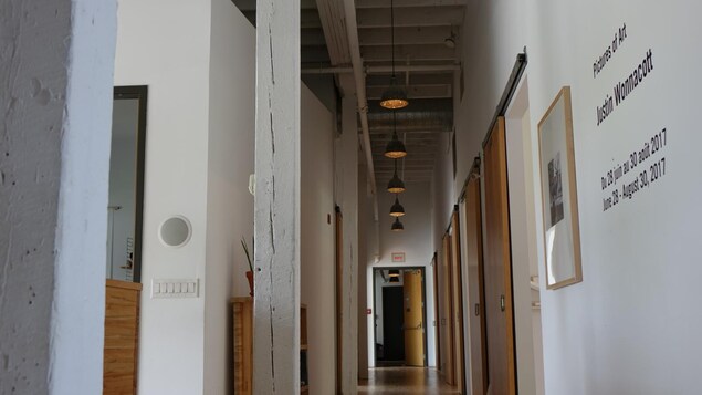 Image montrant un couloir avec plusieurs portes, un plafond haut et des et des luminaires industriels. Image montrant un couloir avec plusieurs portes, un plafond haut et des et des luminaires industriels.