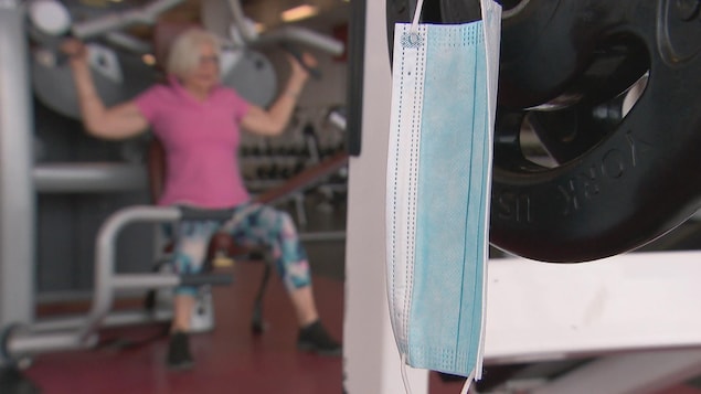 Un masque accroché à un poids, dans un centre de conditionnement physique. En arrière-plan, sur une machine de musculation, on voit une femme en train de s'entraîner.