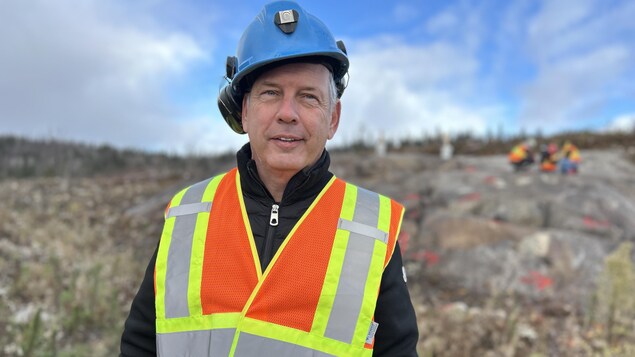 Un homme portant un casque et une veste de visibilité sourit à la caméra devant un paysage rocheux.