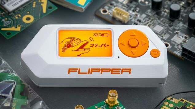Un petit appareil blanc arborant le nom Flipper avec un écran orange sur une table grise entourée de circuits électroniques.