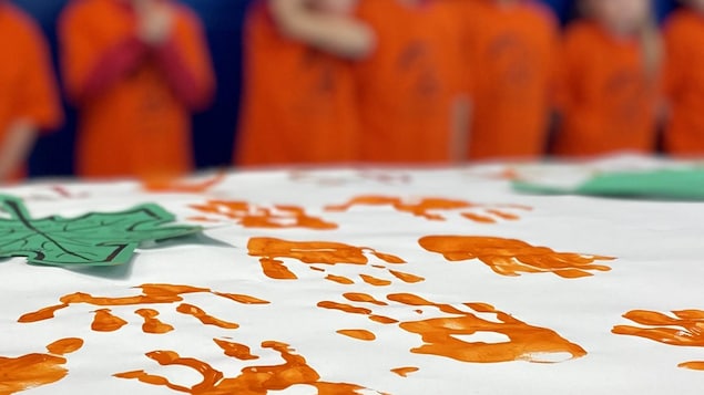 Une bannière contenant des traces de main orange est déposée sur une table. Derrière, des silhouettes d'enfants floues.