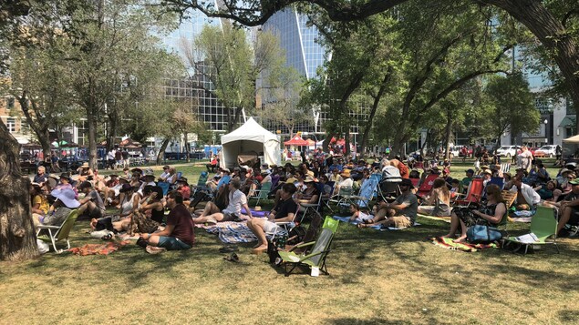 Une centaine de personnes assises sur des chaises de fortune ou sur l'herbe, dans un parc. Une centaine de personnes assises sur des chaises de fortune ou sur l'herbe, dans un parc.