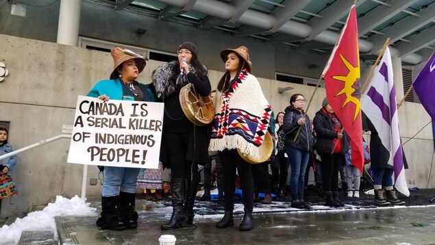 Trois femmes sur le devant de la scène, dont une est au micro et trois autres tiennent des drapeaux derrière. Trois femmes sur le devant de la scène, dont une est au micro et trois autres tiennent des drapeaux derrière.