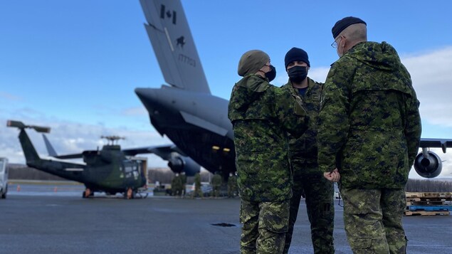 Trois militaires en uniforme parlent en cercle sur un tarmac. On voit des hélicoptères et des avions derrière eux.