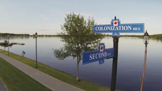 Sur le bord d'une rivière, un chemin avec une pancarte où on voit le nom des rues Colonisation et Second street.