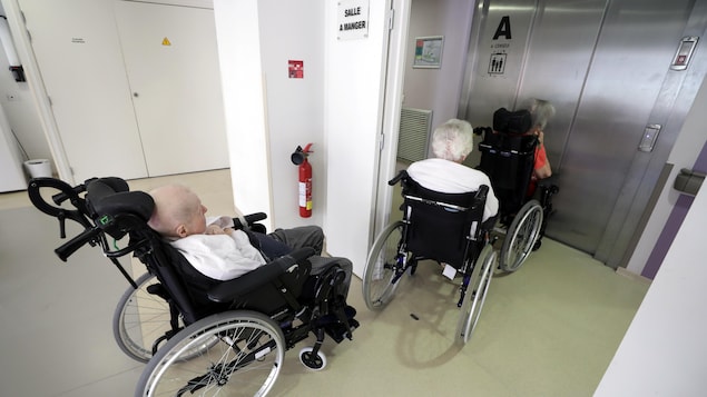 Trois personnes âgées dans leur fauteuil roulant, en rang, devant la porte fermée d'un ascenseur.