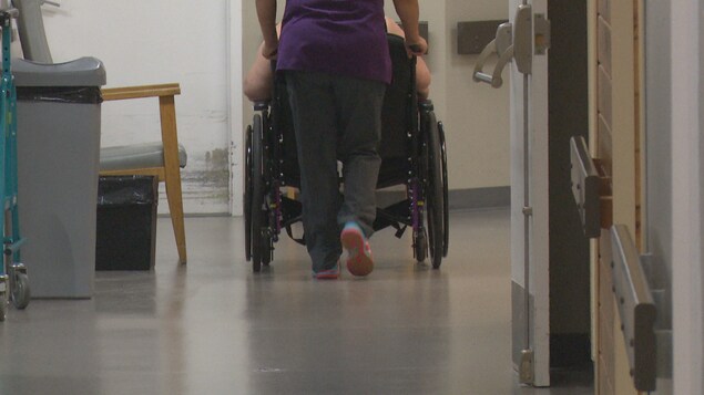 Une préposée aide un résident à se déplacer en fauteuil roulant dans un couloir du foyer de soins.