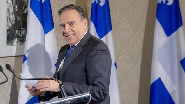 François Legault tient un document en regardant la caméra, derrière un podium.