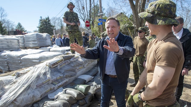 Un homme qui porte des jeans et un manteau discute avec des militaires qui remplissent des sacs de sable.