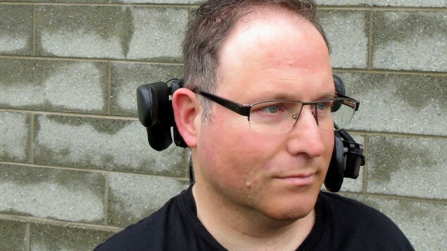 Portrait en couleur de François Marcotte, en extérieur, de trois quarts, devant un mur de blocs de béton. Il porte des lunettes, un t-shirt noir et sa tête est soutenue par l'appui-tête de son fauteuil roulant.