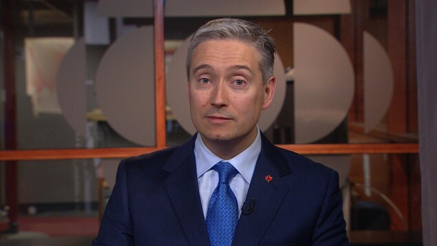 Un homme portant un veston et une cravate regarde droit devant lui. Il est assis devant une vitre sur laquelle se trouve le logo de Radio-Canada.