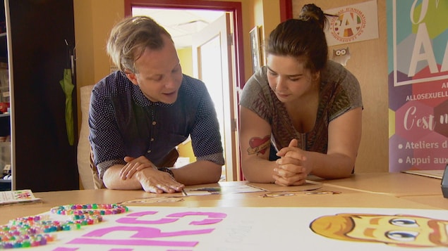 Casey Edmunds et Natacha Coones préparent les affiches pour le défilé de la Fierté, ils sont penchés au-dessus d'une table remplie de dessins multicolores.