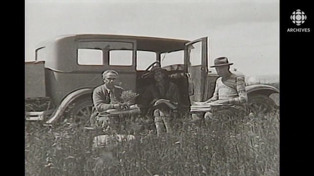 Frère Marie-Victorin et deux membres de son équipe assis dans un champ près d'une voiture, vers 1920.