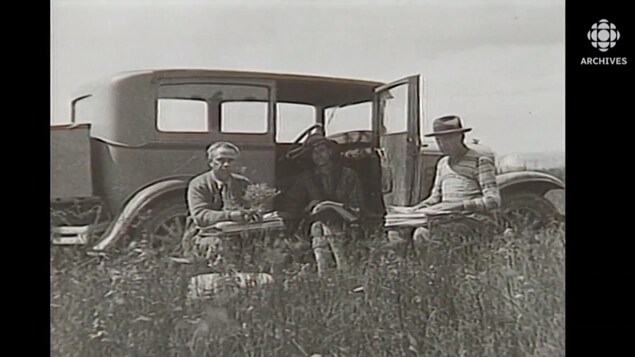 Frère Marie-Victorin et deux membres de son équipe assis dans un champ près d'une voiture, vers 1920.
