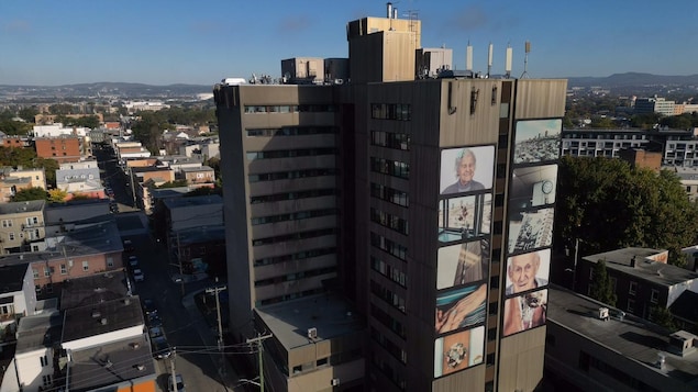 Vue aérienne d'un imposant bâtiment à étages. De grandes photos en couleur ont été collées sur une des façades.