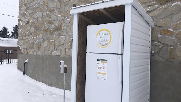 À l'extérieur, en hiver, un frigo est protégé par un abri, tout près du mur d'un bâtiment. 