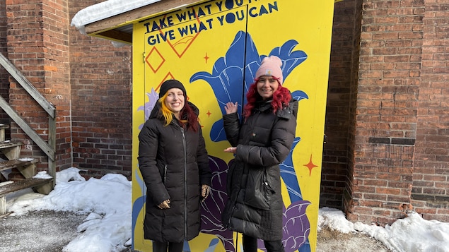 Alex Allaby et Ana Zendejas devant l’abri d’un des frigos communautaires de Toronto. L’abri est peinturé en jaune avec des fleurs bleu et mauve.