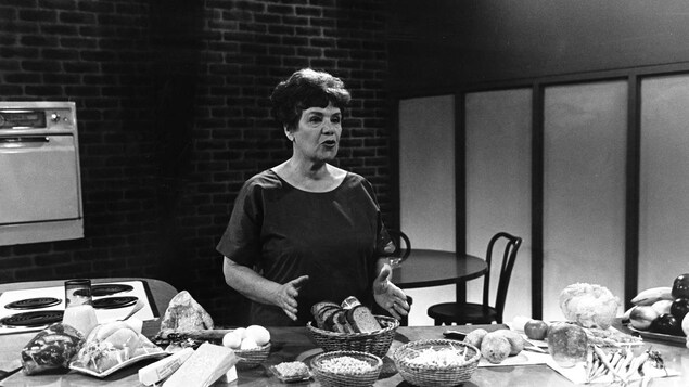 Dans un studio de télévision, la cuisinière Jehane Benoit, debout derrière un comptoir de cuisine, présente divers aliments, dont des fromages, du pain et des légumes.