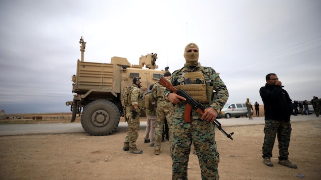 Un soldat masqué porte une arme au premier plan. Au second est posté un véhicule blindé.